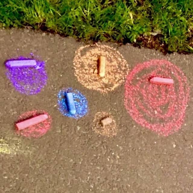 colourful chalk dots with sticks of pavement chalk sitting on them. Pathway and grass.